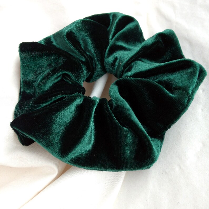 Puede incluir: Una goma para el pelo de terciopelo verde esmeralda oscuro. La tela es suave y afelpada, con textura y pliegues visibles. La goma es un accesorio para el cabello, dise&ntilde;ado para sujetar el pelo en una coleta o mo&ntilde;o.