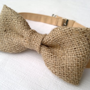 May include: A rustic bow tie made of woven burlap fabric. The bow tie is a light brown color with a beige adjustable neckband and a metal clasp. The bow tie is a classic accessory for formal or casual wear.