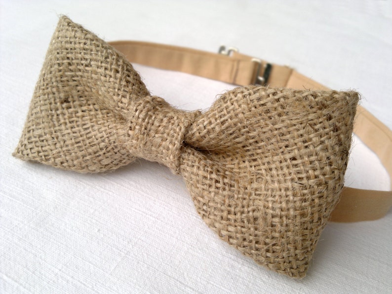 May include: A rustic bow tie made of woven burlap fabric. The bow tie is a light brown color with a beige adjustable neckband and a metal clasp. The bow tie is a classic accessory for formal or casual wear.