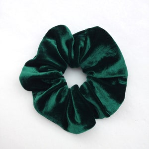 Puede incluir: Una goma para el pelo de terciopelo verde esmeralda oscuro, un accesorio para el cabello, sobre un fondo blanco. La goma es redonda con un dise&ntilde;o fruncido, mostrando la textura afelpada de la tela. El color es rico y saturado.