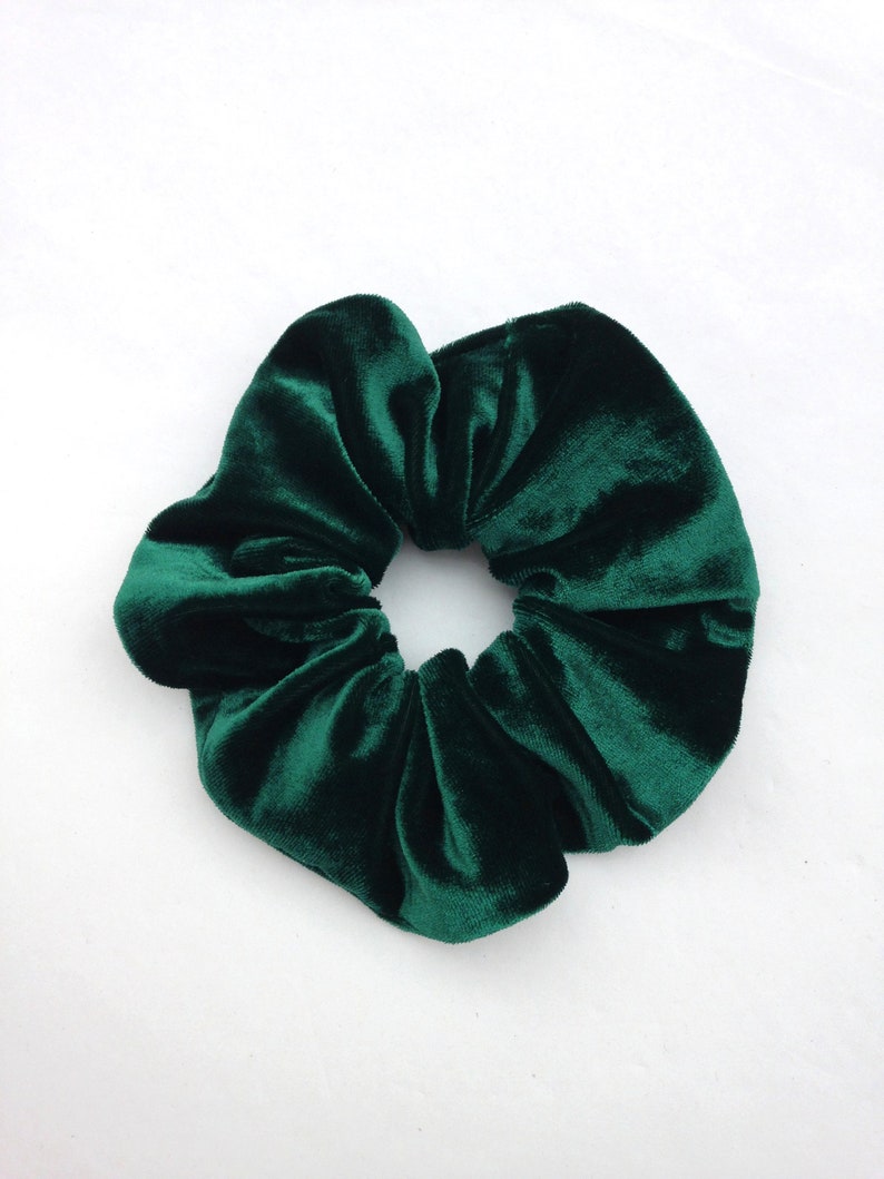 Puede incluir: Una goma para el pelo de terciopelo verde esmeralda oscuro, un accesorio para el cabello, sobre un fondo blanco. La goma es redonda con un dise&ntilde;o fruncido, mostrando la textura afelpada de la tela. El color es rico y saturado.