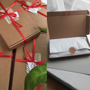 Puede incluir: Varias cajas de cart&oacute;n marr&oacute;n atadas con cinta roja y detalles de arpillera. Una caja est&aacute; abierta, revelando papel de seda blanco y una pegatina redonda que dice "Thank you!". Las cajas son probablemente para regalos o embalaje.