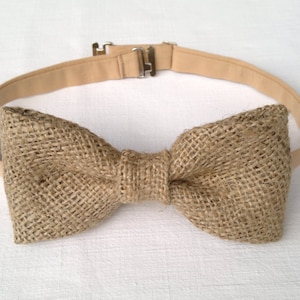 May include: A rustic bow tie made of woven burlap fabric. The bow tie is a natural tan color and is attached to a matching adjustable strap with a metal clasp. The bow tie is centered on a white background.