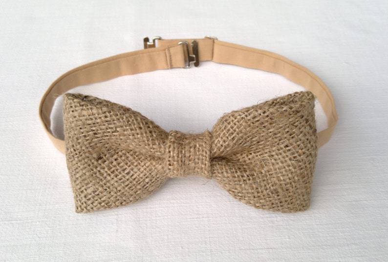May include: A rustic bow tie made of woven burlap fabric. The bow tie is a natural tan color and is attached to a matching adjustable strap with a metal clasp. The bow tie is centered on a white background.