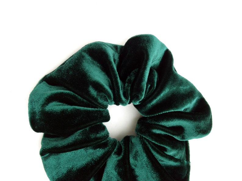 Puede incluir: Una goma para el pelo de terciopelo verde esmeralda oscuro, un accesorio para el cabello, sobre un fondo blanco. La goma para el pelo est&aacute; fruncida y tiene una textura suave y afelpada. La tela refleja la luz, resaltando su rico color y creando sombras.
