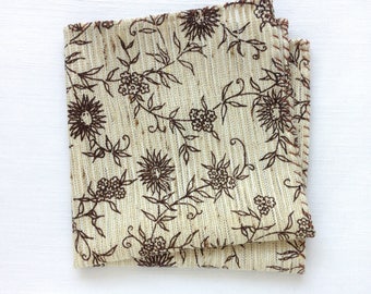 May include: Folded beige cloth napkin with a brown floral pattern. The design features stylized flowers and vines. The napkin is made of a textured fabric and has a folded edge.