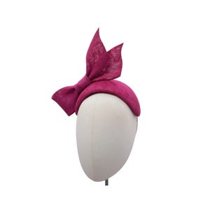 May include: A magenta fabric hat with a large bow on top. The hat is on a white mannequin head.