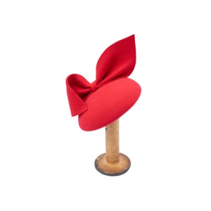 May include: A red felt hat with a large bow on top. The hat is displayed on a wooden stand.