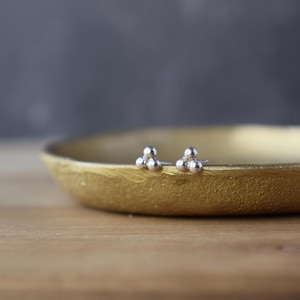 May include: A pair of silver stud earrings, each featuring three small, spherical beads clustered together. The earrings are displayed on a gold-colored, shallow dish, set against a blurred background.