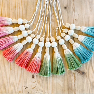 May include: A collection of decorative tassels with wooden beads and ombre-dyed fringe. The tassels are in shades of pink, green, and teal, attached to white cords. The beads are natural wood color and geometric shapes.