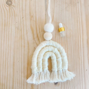 May include: White macrame rainbow wall hanging with wooden beads and a small glass bottle with a gold cap.