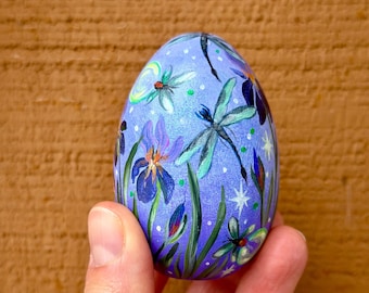 Op de afbeelding: Een handbeschilderd paasei met een paars, blauw en groen ontwerp. Het ei heeft libellen, bloemen en sterren.