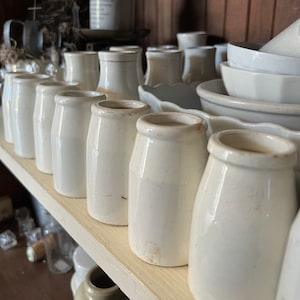 May include: A shelf filled with white ceramic milk bottles, some with a slight yellow tint. The bottles are arranged in a row, with the largest bottle at the back and the smallest bottle at the front. The bottles are all in focus, and the background is blurred.