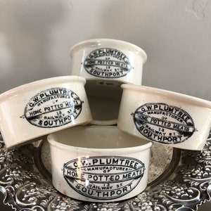 May include: Four white ceramic jars with black lettering. The jars are labeled "C.W. Plumtree, Manufacturer of Home Potted Meats, 13 Railway St, Southport".