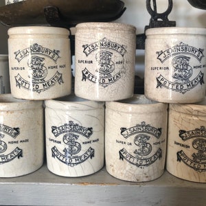 May include: A group of vintage, cream-coloured ceramic pots, each bearing the black text "J. Sainsbury's Potted Meats". The cylindrical pots show signs of age, with cracked glaze. The pots are displayed on a shelf, evoking a sense of history and nostalgia.