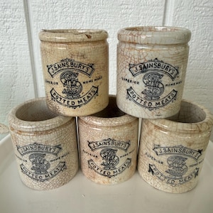 May include: A collection of five antique, cylindrical J. Sainsbury's Potted Meats jars. The jars are off-white with a cracked glaze and black lettering. Each jar has the text "J. Sainsbury's Superior Potted Meats Home Made".