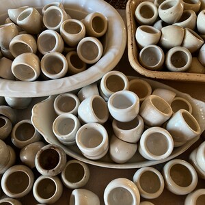 Peut inclure: Une collection de petites tasses en céramique blanc cassé, peut-être pour des sauces ou de petites portions. Les tasses sont disposées dans divers récipients, dont un plat ovale blanc, un plateau rectangulaire et un panier en fil de fer, créant une esthétique rustique.