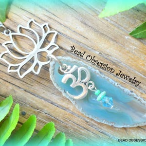May include: A silver charm necklace with a lotus flower pendant and a blue agate slice with a silver Om symbol charm. The agate slice has a white border and a light blue center. The necklace is on a wooden surface with green leaves.