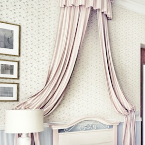 Könnte beinhalten: Ein Schlafzimmer mit einem Himmelbett. Das Bett hat ein weißes Kopfteil und eine blaue Bettdecke mit Blumenmuster. Das Himmelbett besteht aus rosa-weiß gestreiftem Stoff. Eine weiße Lampe steht auf einem passenden Nachttisch.
