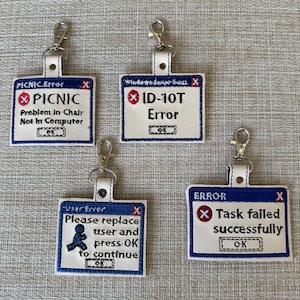 May include: Four white fabric keychains with blue borders and red X marks. Each keychain features a different humorous computer error message. The messages are "PICNIC Problem in Chair Not In Computer", "ID-10T Error", "Please replace user and press OK to continue", and "Task failed successfully".