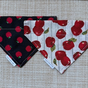 May include: Two triangular pet bandanas featuring apple patterns. One bandana is black with a red apple print, and the other is a light gray wood-grain background with red apples and green leaves. Each has a white tag reading Penny's Paw-sh Pets.