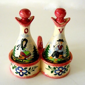 May include: A ceramic oil and vinegar set with a handle, featuring two bottles with red stoppers. Each bottle has a painted scene of people in traditional clothing. The base is decorated with blue flowers and green leaves.