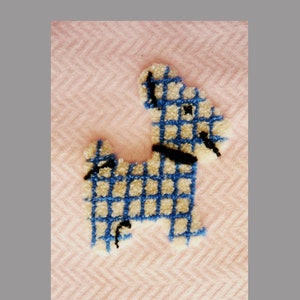 May include: A handmade, embroidered dog design in blue and cream, with a black collar and accents. The dog is set against a pink background with a subtle herringbone pattern. The craftwork is likely a patch or applique.