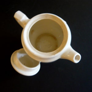 May include: Overhead view of a cream-colored ceramic teapot with its lid slightly ajar. The teapot has a spout, handle, and a round opening. The interior shows signs of use. The background is a dark surface.