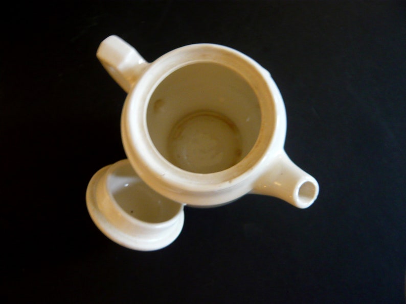 May include: Overhead view of a cream-colored ceramic teapot with its lid slightly ajar. The teapot has a spout, handle, and a round opening. The interior shows signs of use. The background is a dark surface.