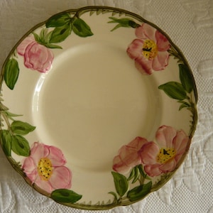 May include: A white ceramic plate with a floral design featuring pink roses and green leaves. The plate has a scalloped edge.