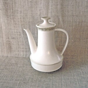 May include: A white ceramic coffee pot with a lid and a handle. The pot has a spout and a decorative band around the top. The base of the pot has vertical ridges. The background is a neutral, textured fabric.