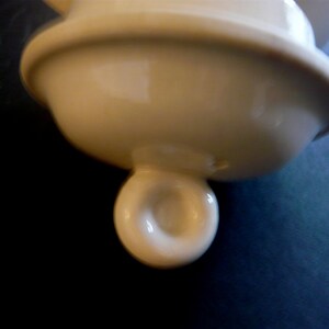 May include: Cream-colored ceramic lid with a rounded top and a spherical handle. The lid has a smooth, glossy finish and appears to be part of a larger ceramic piece. The handle has a hole in the center.