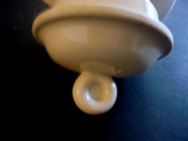 May include: Cream-colored ceramic lid with a rounded top and a spherical handle. The lid has a smooth, glossy finish and appears to be part of a larger ceramic piece. The handle has a hole in the center.