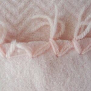 May include: Close-up of a light pink fabric with a textured chevron pattern. The edge is finished with a decorative stitch in the same color, and loose threads extend from the edge.