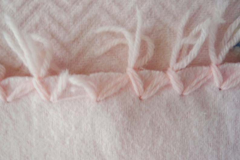 May include: Close-up of a light pink fabric with a textured chevron pattern. The edge is finished with a decorative stitch in the same color, and loose threads extend from the edge.