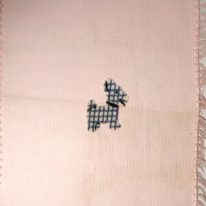 May include: A light pink square blanket with a blue and white checkered dog applique. The blanket has a fringed edge in a matching pink color. The fabric has a subtle chevron pattern.