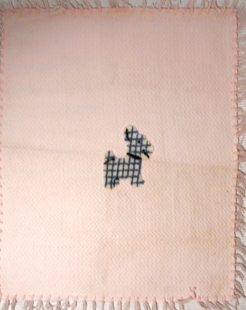 May include: A light pink square blanket with a blue and white checkered dog applique. The blanket has a fringed edge in a matching pink color. The fabric has a subtle chevron pattern.