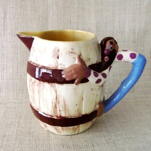 May include: A ceramic pitcher with a barrel design, featuring a figure with a brown beard and polka-dot shirt. The handle is blue, and the pitcher has a spout. The barrel is cream-colored with brown bands.