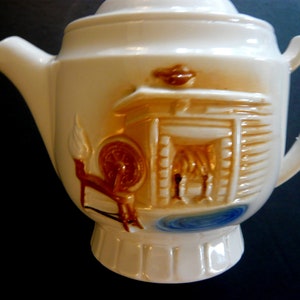 May include: Cream-colored ceramic teapot with a lid and handle. The teapot features a raised relief design of a spinning wheel, a cabin, and a small pool of water, all in shades of brown and blue.