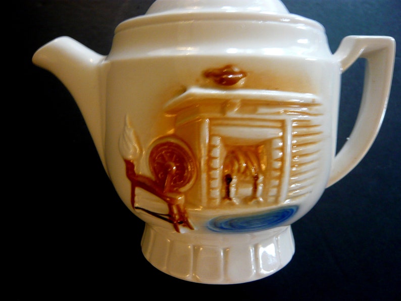 May include: Cream-colored ceramic teapot with a lid and handle. The teapot features a raised relief design of a spinning wheel, a cabin, and a small pool of water, all in shades of brown and blue.