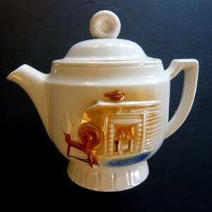 May include: Cream-colored ceramic teapot with a lid and handle. The teapot features a raised relief design of a fireplace, spinning wheel, and blue accents. The spout and handle are smooth, while the body has a textured appearance.