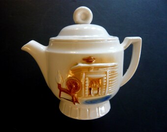 May include: Cream-colored ceramic teapot with a lid and handle. The teapot features a raised relief design of a fireplace, spinning wheel, and blue accents. The spout and handle are smooth, while the body has a textured appearance.