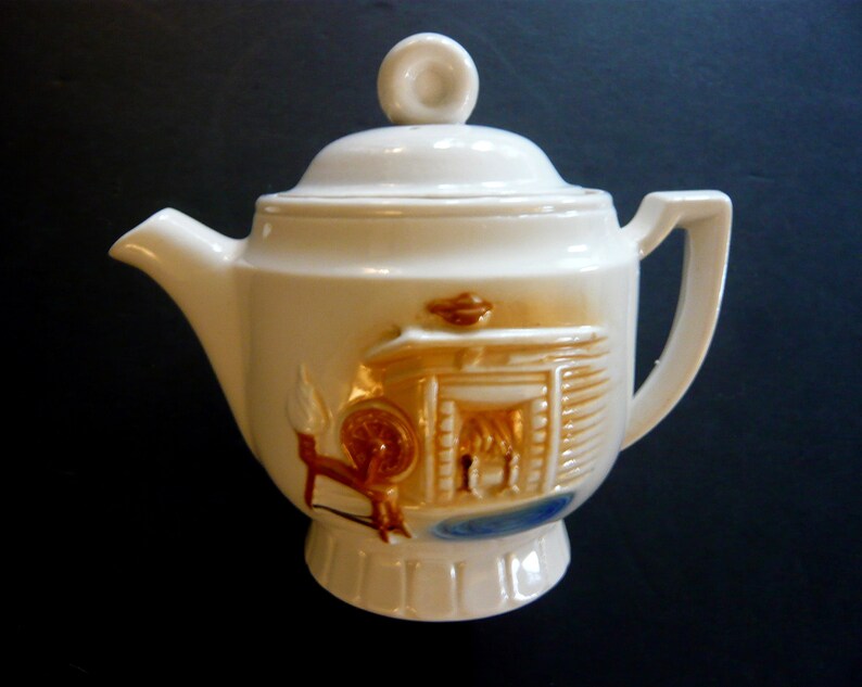 May include: Cream-colored ceramic teapot with a lid and handle. The teapot features a raised relief design of a fireplace, spinning wheel, and blue accents. The spout and handle are smooth, while the body has a textured appearance.