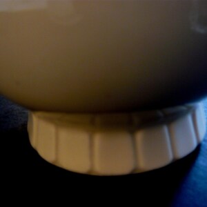 May include: A cream-colored ceramic bowl with a rounded body and a fluted base. The bowl's surface is smooth and glossy, and the base has a series of vertical indentations. The bowl is likely for serving food.