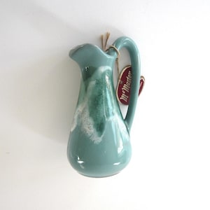 May include: A teal ceramic pitcher with a curved handle and a ruffled rim. The pitcher has a glossy finish with white and green accents. A red tag with the text "McMaster" is attached to the handle with twine. The pitcher is a decorative item.
