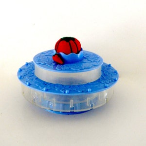 May include: A blue and clear plastic sewing kit with a red tomato-shaped pin cushion on top. The kit has a two-tiered design with floral detailing and a clear, cylindrical container.