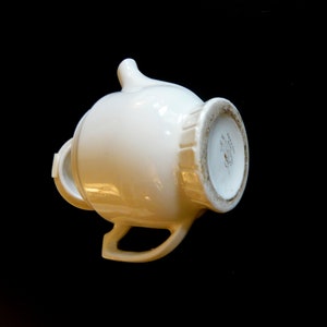 May include: A white ceramic creamer with a handle and spout, set against a black background. The creamer has a rounded body and a decorative rim. The bottom of the creamer has a detailed design.
