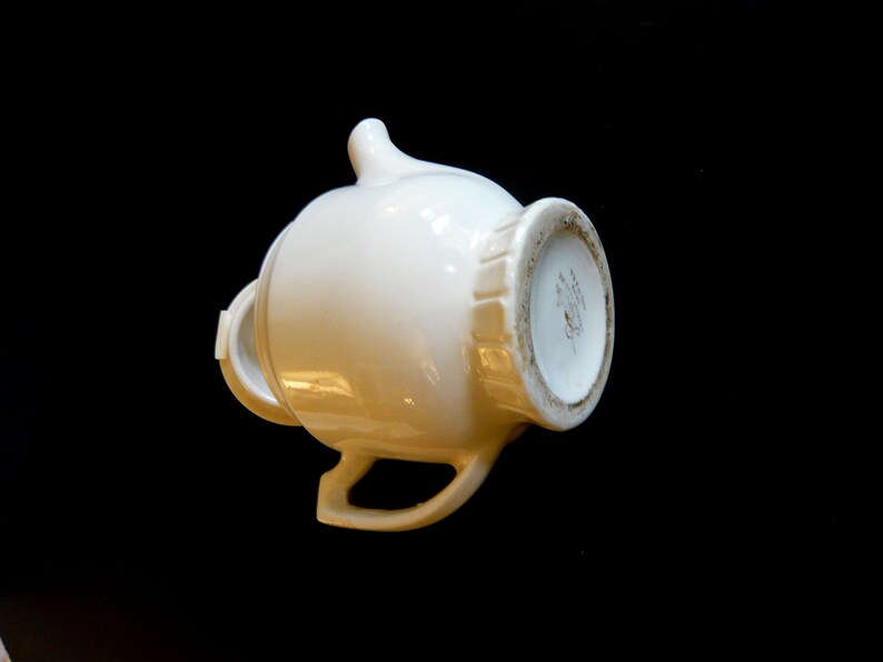 May include: A white ceramic creamer with a handle and spout, set against a black background. The creamer has a rounded body and a decorative rim. The bottom of the creamer has a detailed design.