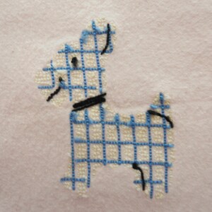 May include: Embroidered dog design in blue and black on a light pink fabric. The dog is created with a grid pattern, with a black collar and details. The background is a soft, solid pink.