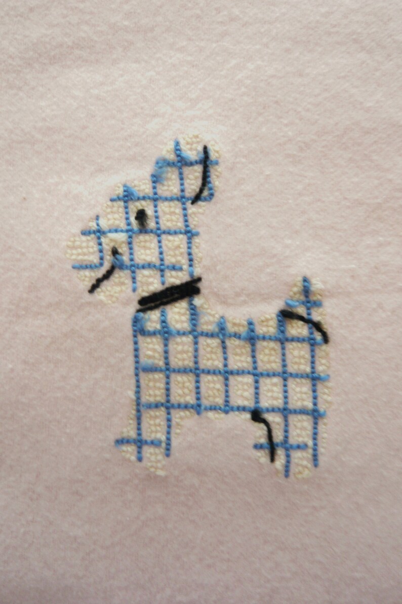 May include: Embroidered dog design in blue and black on a light pink fabric. The dog is created with a grid pattern, with a black collar and details. The background is a soft, solid pink.
